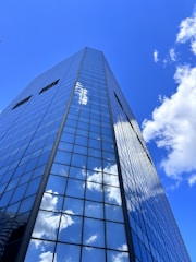 A tall, modern skyscraper with reflective glass panels stretches upwards against a bright blue sky. Fluffy white clouds are mirrored across the building's facade, creating a seamless blend between architecture and nature.