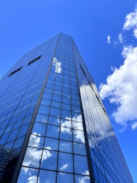 A tall, modern skyscraper with reflective glass panels stretches upwards against a bright blue sky. Fluffy white clouds are mirrored across the building's facade, creating a seamless blend between architecture and nature.
