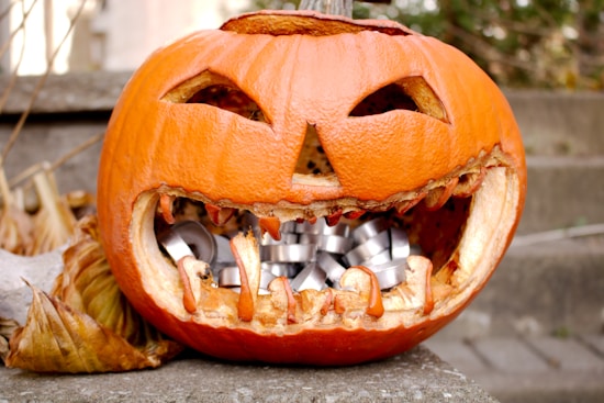A carved pumpkin with a menacing expression featuring triangular eyes and a jagged mouth. The interior is hollowed out and filled with shiny silver objects. Brown, dried leaves surround the pumpkin, hinting at an autumn setting. The background shows stone steps.