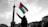 A person stands holding a Palestinian flag prominently against a cloudy sky. In the background, there is a tall column with a statue on top. People are gathered, with some signs visible advocating for freedom and resistance.