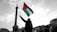 A person stands holding a Palestinian flag prominently against a cloudy sky. In the background, there is a tall column with a statue on top. People are gathered, with some signs visible advocating for freedom and resistance.