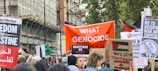 A protest scene with numerous people holding signs and banners. Prominent messages include demands for freedom and sanctions, and opposition to genocide and occupation. The crowd is diverse, and some people hold Palestinian flags. The background includes buildings and trees, suggesting an urban environment.