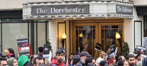 A crowd of people gathered in front of a building with a sign that reads 'The Dorchester The Ballroom'. Many individuals are holding signs and banners with messages such as 'Stop Genocide'. The atmosphere appears to be that of a protest or demonstration.