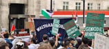 A crowd of people is gathered for a protest in an urban setting, holding various signs and banners advocating against Israeli actions and calling for the end of apartheid and genocide. The Palestinian flag is visible, and participants seem engaged and focused.