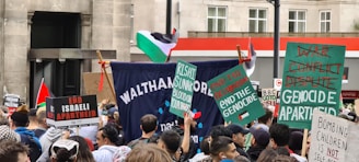 A crowd of people is gathered for a protest in an urban setting, holding various signs and banners advocating against Israeli actions and calling for the end of apartheid and genocide. The Palestinian flag is visible, and participants seem engaged and focused.