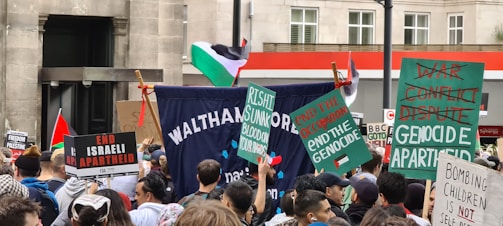A crowd of people is gathered for a protest in an urban setting, holding various signs and banners advocating against Israeli actions and calling for the end of apartheid and genocide. The Palestinian flag is visible, and participants seem engaged and focused.