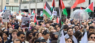 A large crowd of people gathered in a city holding flags, signs, and banners advocating for Palestinian freedom. The group is diverse, with many individuals wearing traditional Palestinian scarves. The Palestinian flags prominently feature red, green, black, and white colors, and several placards convey messages of solidarity and calls for action.