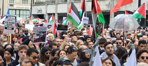 A large crowd of people gathered in a city holding flags, signs, and banners advocating for Palestinian freedom. The group is diverse, with many individuals wearing traditional Palestinian scarves. The Palestinian flags prominently feature red, green, black, and white colors, and several placards convey messages of solidarity and calls for action.