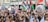 A large crowd of people gathered in a city holding flags, signs, and banners advocating for Palestinian freedom. The group is diverse, with many individuals wearing traditional Palestinian scarves. The Palestinian flags prominently feature red, green, black, and white colors, and several placards convey messages of solidarity and calls for action.