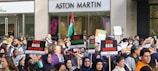 A large group of people is gathered in a protest outside a building with the Aston Martin logo. They are holding signs with messages like 'Stop Gaza Genocide' and 'Free Palestine'. A Palestinian flag is visible among the crowd, and many participants appear passionate and engaged in the demonstration.