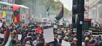 A large crowd of people is gathered in a city street, holding signs and flags with political messages. The scene includes red double-decker buses, and various placards are visible while people demonstrate peacefully. Trees and buildings line the street.