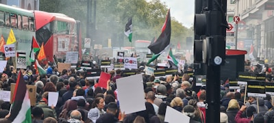 A large crowd of people is gathered in a city street, holding signs and flags with political messages. The scene includes red double-decker buses, and various placards are visible while people demonstrate peacefully. Trees and buildings line the street.