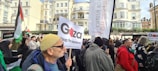 A large group of people are gathered in a city setting, holding signs and flags in a protest. Buildings with traditional architecture are visible in the background. The signs display messages related to Gaza, and many people are wearing winter clothing.