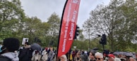 A crowd of people gather outdoors near a tall red Labour Party banner amid greenery and traffic lights. Some attendees hold umbrellas and protest signs.