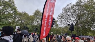 A crowd of people gather outdoors near a tall red Labour Party banner amid greenery and traffic lights. Some attendees hold umbrellas and protest signs.