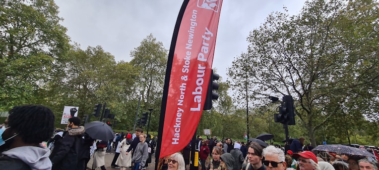 A crowd of people gather outdoors near a tall red Labour Party banner amid greenery and traffic lights. Some attendees hold umbrellas and protest signs.