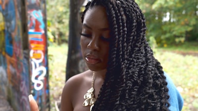 A person with long, braided hair stands near a wall covered with colorful graffiti. The background includes greenery and trees, suggesting an outdoor setting. The person is wearing a necklace with large, metallic shapes.