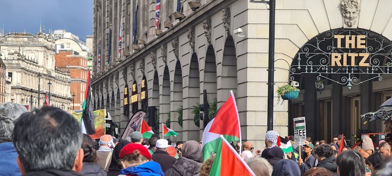 A crowd of people is gathered in front of The Ritz hotel. Many individuals are holding and waving red, green, black, and white flags. The hotel building features a classical facade with arched windows and flags displayed above. In the background, there are other buildings and a cloudy sky.