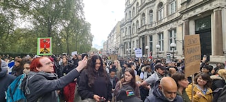 A large group of people participates in a protest march along a city street lined with old, ornate buildings. Many individuals hold signs with various messages, and some carry flags. The crowd includes a diverse mix of individuals, and the atmosphere is busy and engaged.