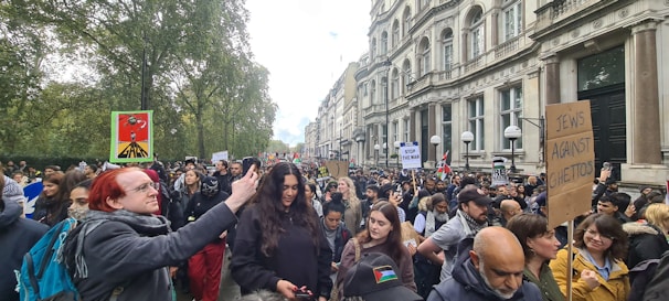 A large group of people participates in a protest march along a city street lined with old, ornate buildings. Many individuals hold signs with various messages, and some carry flags. The crowd includes a diverse mix of individuals, and the atmosphere is busy and engaged.