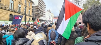 A large crowd of people gathered for a protest or demonstration. Various flags and signs are visible, including a prominent flag featuring red, green, black, and white colors. The surrounding buildings and construction scaffolding indicate an urban setting.