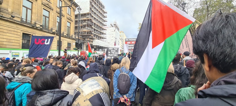 A large crowd of people gathered for a protest or demonstration. Various flags and signs are visible, including a prominent flag featuring red, green, black, and white colors. The surrounding buildings and construction scaffolding indicate an urban setting.