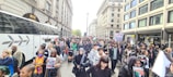 A large group of people are participating in a street protest. Many are holding signs and flags, and the crowd fills the city street, bordered by tall commercial buildings. A white coach bus is parked on the side. The atmosphere is bustling and diverse, with individuals of various ages and backgrounds involved.