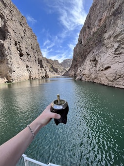 A hand holding a traditional mate cup above a body of water, with steep rocky cliffs on either side. The sky is clear with some clouds, and the scene conveys a sense of tranquility and natural beauty.