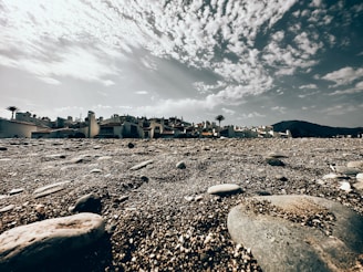 A rocky beach foreground with smooth stones scattered across pebble-covered ground. In the background, a cluster of white, Spanish-style houses with red-tiled roofs and a few palm trees. The scene is under an expansive sky filled with scattered, fluffy clouds, creating a dramatic atmosphere.