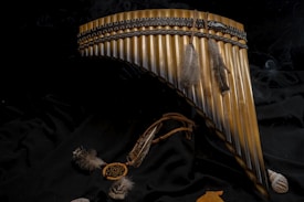 A pan flute adorned with two feathers is placed on a dark fabric. Surrounding the flute are a small dreamcatcher-like ornament and a few seashells. The arrangement gives a sense of cultural and natural elements coming together.