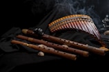 Several wooden wind instruments are arranged on a dark fabric background. The collection includes panpipes and various flutes with intricate designs. A subtle wisp of smoke adds to the atmosphere.