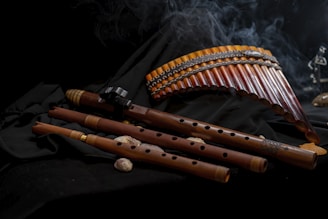 Several wooden wind instruments are arranged on a dark fabric background. The collection includes panpipes and various flutes with intricate designs. A subtle wisp of smoke adds to the atmosphere.