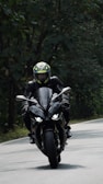 A motorcyclist wearing a helmet and leather gear rides a sleek, black sport motorcycle on a winding road surrounded by dense greenery.