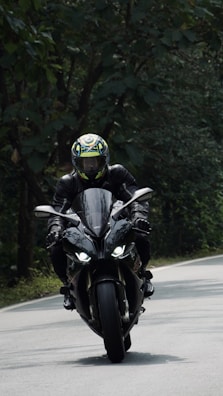 A motorcyclist wearing a helmet and leather gear rides a sleek, black sport motorcycle on a winding road surrounded by dense greenery.