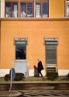 An elderly man walking on a sidewalk in front of an orange building with large windows and two signs advertising art-related services in a foreign language. He casts a shadow on the wall, and small trimmed bushes line the pathway. A bent traffic sign is visible at the edge of the walkway.