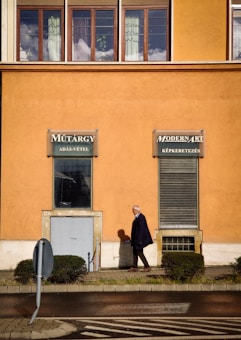 An elderly man walking on a sidewalk in front of an orange building with large windows and two signs advertising art-related services in a foreign language. He casts a shadow on the wall, and small trimmed bushes line the pathway. A bent traffic sign is visible at the edge of the walkway.