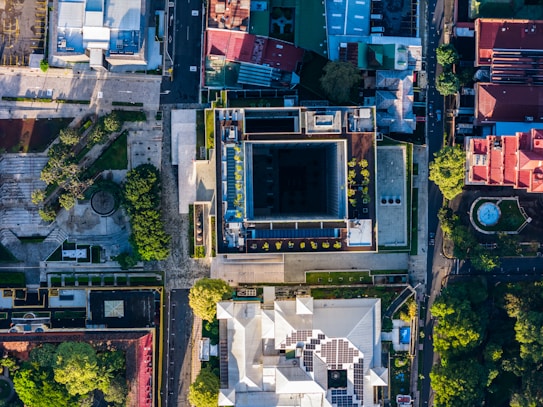 An aerial view of an urban area showcasing several buildings with distinct architectural designs. The image captures a variety of rooftops, including some with solar panels and vegetation. Streets and paths are visible, with green trees interspersed among the structures, adding a touch of nature amidst the urban environment.