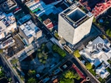 An aerial view of an urban area featuring a large, modern rectangular skyscraper surrounded by smaller buildings and streets. Solar panels are visible on one of the rooftops, and various trees and greenery are scattered throughout the scene. The shadows and lighting suggest the image was taken during late afternoon or early morning.