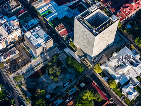 An aerial view of an urban area featuring a large, modern rectangular skyscraper surrounded by smaller buildings and streets. Solar panels are visible on one of the rooftops, and various trees and greenery are scattered throughout the scene. The shadows and lighting suggest the image was taken during late afternoon or early morning.