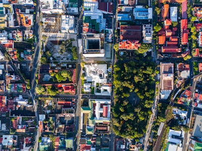 An aerial view of a city landscape with a grid-like pattern of streets. Buildings of various sizes and shapes are interspersed with green spaces and tree-lined areas. The rooftops feature a mix of colors, including red, green, and white. A large park or forested area is visible in the center-right area, providing a contrast to the urban layout.