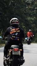 Two motorcyclists are riding along a tree-lined road. The person nearest to the camera wears a colorful helmet and a black shirt with the phrase 'Live it now!' printed on it with playful graphics. The other rider in the distance wears a red jacket. The setting is outdoors with green foliage on both sides of the road.