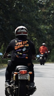 Two motorcyclists are riding along a tree-lined road. The person nearest to the camera wears a colorful helmet and a black shirt with the phrase 'Live it now!' printed on it with playful graphics. The other rider in the distance wears a red jacket. The setting is outdoors with green foliage on both sides of the road.