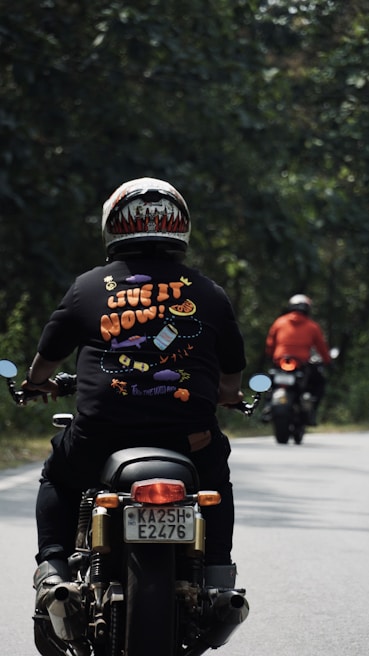 Two motorcyclists are riding along a tree-lined road. The person nearest to the camera wears a colorful helmet and a black shirt with the phrase 'Live it now!' printed on it with playful graphics. The other rider in the distance wears a red jacket. The setting is outdoors with green foliage on both sides of the road.