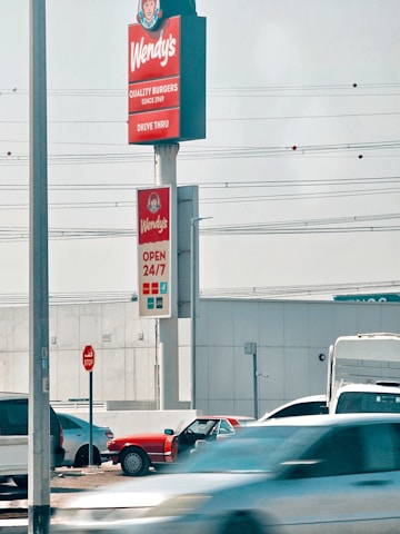 A Wendy's sign displaying slogans like 'QUALITY BURGERS SINCE 1969' and 'OPEN 24/7' is prominently shown. Several cars are parked nearby, with some moving in front of the sign. A stop sign is visible on the left side.
