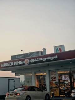 A Burger King fast food restaurant storefront is visible with its recognizable logo and red signage. The front of the building features glass doors and windows, displaying promotional posters for deals and menus. A parked taxi is in front of the restaurant, with a visible license plate. The top right corner of the image reveals another building or structure with signage, suggesting a commercial area. The sky is overcast, giving an early morning or late afternoon ambiance.