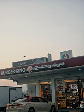 A Burger King fast food restaurant storefront is visible with its recognizable logo and red signage. The front of the building features glass doors and windows, displaying promotional posters for deals and menus. A parked taxi is in front of the restaurant, with a visible license plate. The top right corner of the image reveals another building or structure with signage, suggesting a commercial area. The sky is overcast, giving an early morning or late afternoon ambiance.