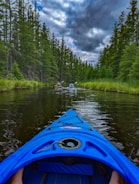 A vibrant blue kayak navigates through a calm, narrow waterway surrounded by dense green forest. The sky above is partly cloudy, contributing to a serene yet slightly overcast atmosphere. Two additional kayakers can be seen ahead, creating a sense of adventure and exploration.