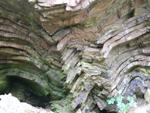Layers of rock are stacked unevenly, with a combination of horizontal and wavy patterns. The rock surface appears to have a variety of textures, with some areas covered in patches of green moss or lichen. The scene suggests a geological formation, possibly formed by natural forces over time.