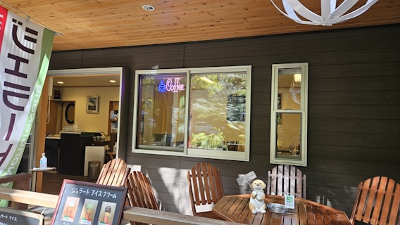 A cozy café setting with a dark exterior wall, featuring a wooden ceiling and Adirondack chairs around a small table. Inside, the café is visible through a large window, illuminated by a blue neon 'Coffee' sign. A small plush toy dog sits on one of the chairs next to an ice cream menu board. There is a colorful flag on the left and a modern light fixture hanging from the ceiling.