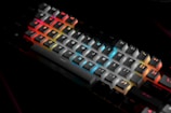 A mechanical keyboard with RGB backlighting, featuring keys in shades of red, blue, orange, and yellow, creating a vibrant, colorful effect against a dark background. Each key is individually lit, highlighting the letters and symbols prominently.